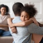 Young african american man holding, embracing, comforting smiling happy calm black cute kid daughter, blurred mother sitting on couch on background, loving supporting family concept.